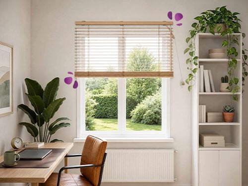 Eine Homeoffice-Ecke mit einer Holzjalousie &uuml;ber einem gro&szlig;en Fenster. Sie filtert das Tageslicht sanft und bietet Blick auf einen gr&uuml;nen Garten. Darunter stehen ein Holzschreibtisch und ein Lederstuhl. Eine hohe Pflanze und wei&szlig;e offene Regale mit kleinen Kakteen auf beiden Seiten schaffen einen ruhigen, luftigen Arbeitsplatz.