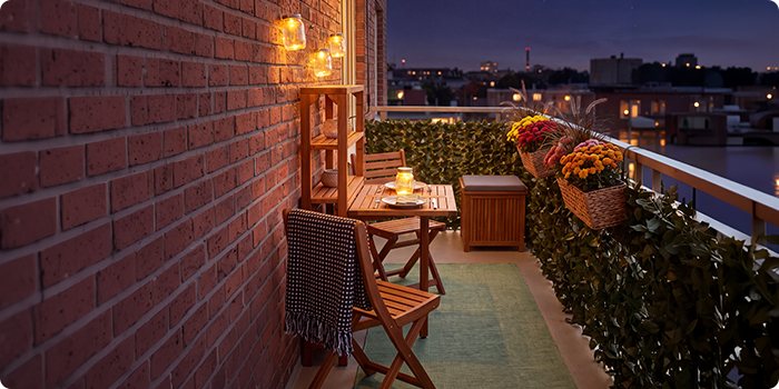 Kleiner Balkon mit Holzstühlen und Tisch, beleuchtet von Laternen und Kerzen, dekoriert mit Topfblumen und Grünpflanzen, mit Blick auf die Skyline der Stadt bei Nacht.