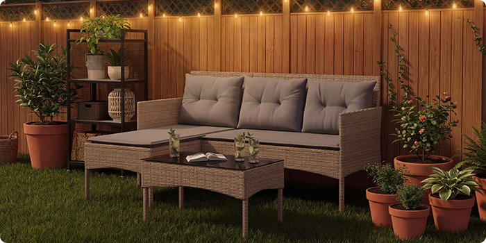 Rattan-Gartenlounge-Set mit grauen Kissen, bestehend aus einem Dreiersitzer, einem Fußhocker und einem Couchtisch auf einem Rasen mit Lichterketten und Topfpflanzen